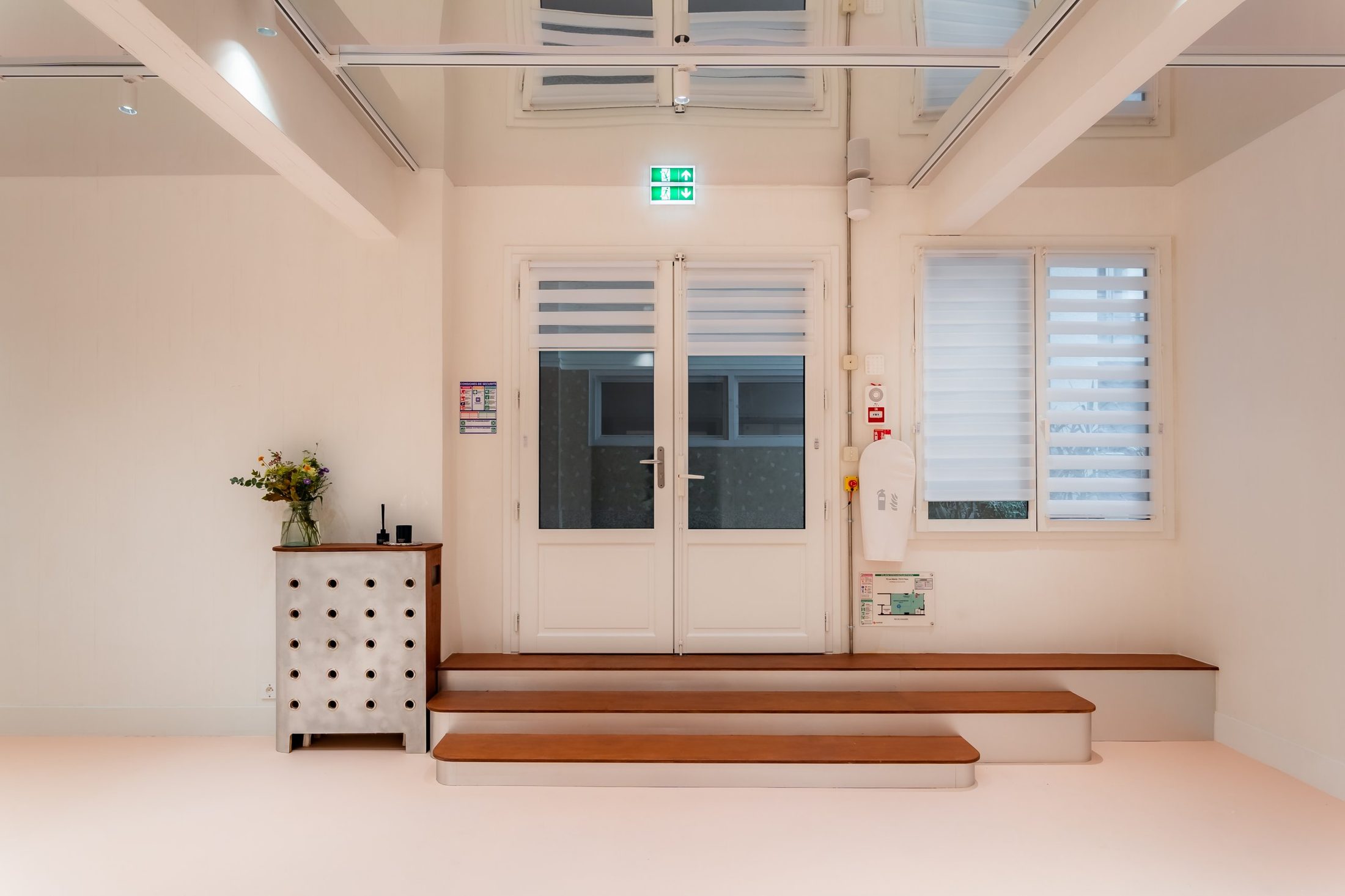 L'entrée de L'Art Room de face : double porte vitrée, trois marches en bois, fenêtre à stores et cache radiateur en aluminium perforé
